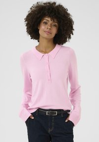 Lichtroze, lange mouwen polo trui met een kraag en een knoopsluiting. Zachte gebreide textuur, gecombineerd met donkere jeans en een zwarte riem.
