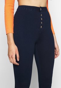 Jeans de tiro alto en azul oscuro, fabricados en denim, que cuentan con un cierre frontal de botones con botones de metal decorativos y un ajuste slim.