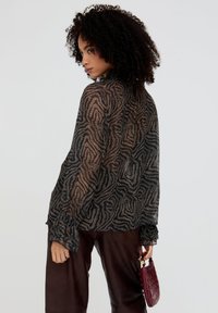 Transparente langärmlige Bluse in Schwarz mit metallisch goldenen Zebramustern, ausgestattet mit gerafften Manschetten und einem hohen Ausschnitt, kombiniert mit braunen Lederhosen.