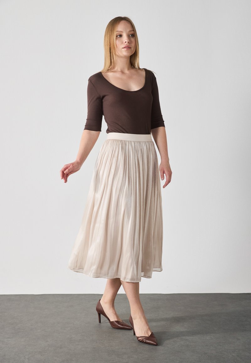 Top marrón ajustado, falda midi plisada color crema y tacones marrones de punta. La falda tiene una textura ligera y fluida y una cinturilla ancha.