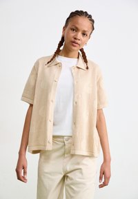 Beige knitted short-sleeve shirt with a button-down front, featuring a textured pattern and a spread collar, worn over a white t-shirt.