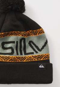 Zwarte gebreide beanie met een fluffy pompon, voorzien van een groene band en een oranje decoratief patroon, waarop het woord "SILV" in het zwart staat.