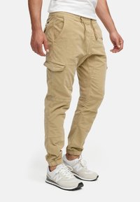 Pantalones cargo caqui con puños elásticos, cintura con cordón, bolsillos laterales y un acabado de mezcla de algodón suave y texturizado. Llevados con zapatillas blancas.