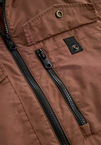 Tissu brun avec deux fermetures éclair verticales noires et un détail de poche à rabat boutonnière sur la surface du vêtement.