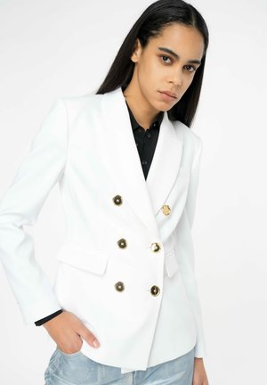 Blazer bianco doppiopetto con bottoni dorati, rever a scollo e tasche laterali. Presenta una texture liscia. Indossato sopra una camicia nera.