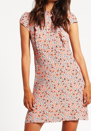 Kobieta w sukience z krótkim rękawem w kolorze brzoskwiniowym z białym nadrukiem kwiatów stokrotki i falbankami wzdłuż brzucha.