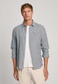 Camisa de cuadros azul y blanca, hecha de tela suave, con un bolsillo en el pecho izquierdo y las mangas enrolladas. Lleva una camiseta blanca debajo.