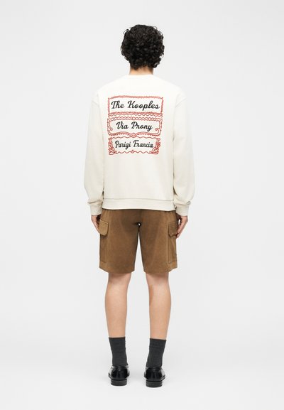 Άτομο στέκεται γυρισμένο με την πλάτη, φοράει κρεμ φούτερ με κείμενο σε κόκκινο πλαίσιο, καφέ cargo σορτς, μαύρα παπούτσια και σκούρες κάλτσες.