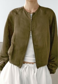 Giacca cropped verde oliva, tessuto leggero, chiusura con zip, scollo rotondo, orlo grezzo, indossata sopra una maglietta bianca a coste e pantaloni bianchi con coulisse.