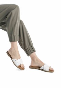 Sandalias deslizantes blancas con tiras cruzadas, suela beige y diseño plano. Combinadas con pantalones grises de longitud hasta el tobillo y con puños, de textura suave.