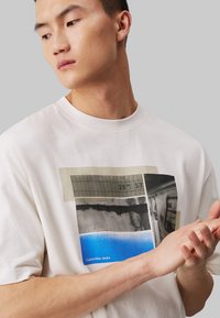 Κρεμ T-shirt με παραλληλόγραμμη εκτύπωση γραφικών που περιλαμβάνει μαύρους, γκρι και μπλε τόνους. Μακριά μανίκια, στρογγυλή λαιμόκοψη, απαλό βαμβακερό ύφασμα.