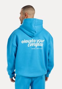 Hoodie en coton bleu avec une capuche à cordon de serrage, comportant un texte blanc au dos qui dit "élever votre quotidien" et des détails de logo.