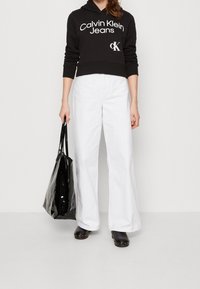 Sudadera negra corta con logo blanco "Calvin Klein Jeans", combinada con pantalones blancos de pierna ancha. La modelo sostiene un bolso tote negro brillante.