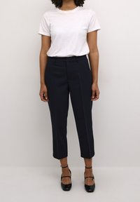 T-shirt blanc à manches courtes associé à un pantalon taille haute bleu marine, avec une coupe ajustée, une texture lisse et une longueur raccourcie. Des talons noirs à bride complètent la tenue.