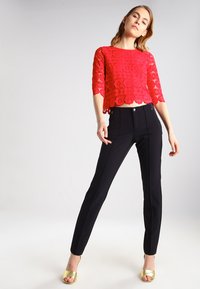 Top corto de encaje rojo con patrones florales, combinado con pantalones negros ajustados de textura suave. La modelo lleva sandalias doradas de tacón.