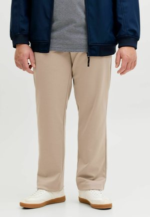 Beige bukser med rette ben, brukt med hvite joggesko, grå skjorte og marineblå zip-up jakke på en person som står mot en ensfarget bakgrunn.