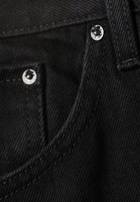 Gros plan sur un jean en denim noir montrant une poche avant, deux rivets métalliques, des coutures et le bord d’un passant de ceinture.