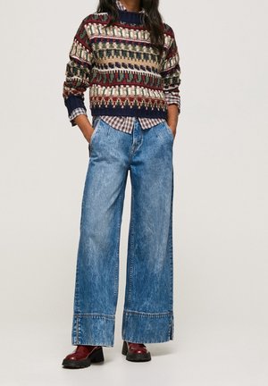 Osoba ubrana w wzorzysty sweter dzianinowy na koszuli w kratę, szerokie niebieskie dżinsy i ciemnoczerwone buty, stojąca z rękami w kieszeniach.