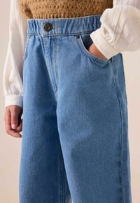 Pantalons en denim bleu clair taille haute avec fermeture à bouton, poches latérales et détails de couture visibles. Le matériau semble robuste et texturé.