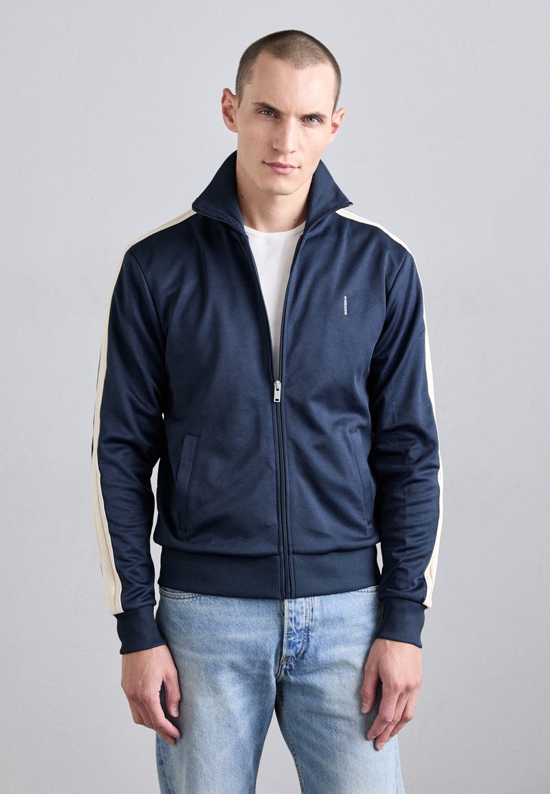 Ung man står klädd i en marinblå zip-up-jacka med vita ränder på ärmarna och ljusblå jeans framför en enkel grå bakgrund.