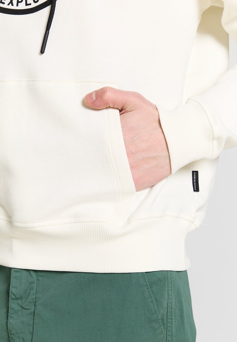 Sudadera con capucha color crema, con un bolsillo frontal y cordones negros. Tejido ligero con puños acanalados, llevada sobre pantalones verdes.