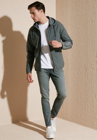 Jachetă ușoară pentru bărbați, cu glugă, de culoare gri închis, asociată cu blugi ajustați gri deschis și adidași albi. Textură netedă, design casual.