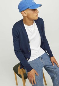 Cardigan blu navy con maniche lunghe, chiusura a bottoni e polsini a costine, indossato sopra una maglietta bianca, abbinato a jeans azzurri chiari.