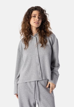 Femme aux longs cheveux bouclés portant un haut gris boutonné ample et un pantalon assorti à cordon, debout devant un fond clair uni.