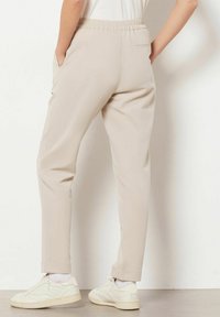 Pantalon beige avec taille élastique et poches latérales, coupe fuselée, revers retroussés, associé à des baskets blanches. Tissu lisse et léger.