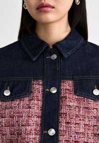 Chaqueta de denim con una sección superior de color azul oscuro, un área inferior texturizada de color rosa en contraste, doble bolsillo en el pecho y botones de hardware plateados.