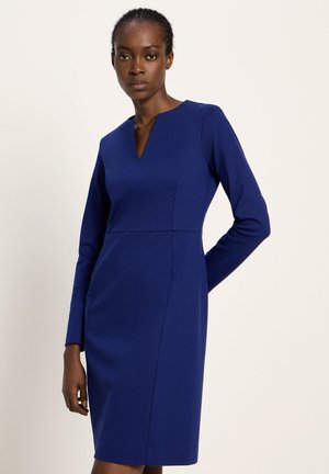 Eine Frau trägt ein eng anliegendes, langärmeliges, knielanges königsblaues Kleid mit einem Ausschnitt und steht vor einem schlichten hellen Hintergrund.