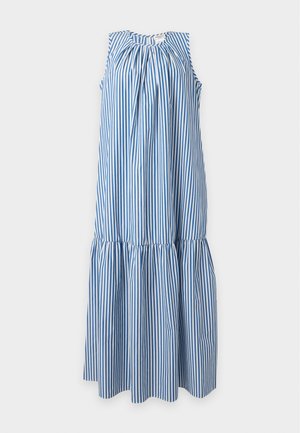 Frau mit langen dunklen Haaren, die ein ärmelloses blau-weiß gestreiftes Maxikleid und braune Stiefeletten trägt, auf weißem Hintergrund.