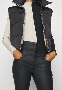 Vadderad svart väst med hög krage över en ribbad beige och svart topp. Svarta högmidjade byxor med fyra knappar och sidfickor.