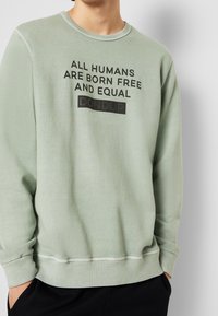 Ljusgrön sweatshirt med svart text som säger "ALLA MÄNNISKOR ÄR FÖDDA FRIA OCH LIKA" och en logotyp. Har ribbade muddar och halsringning.