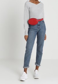 Šedý pletený svetr s výstřihem do V, vysoké modré džíny s ohrnutými manžetami, červená oválná taška kolem pasu a bílé tenisky. Hladké textury a živé barvy.