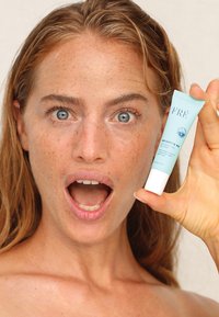 Tubo azzurro chiaro di crema "Brighten Me" di FRÉ con cappuccio bianco, tenuto da una persona con capelli lunghi, che mostra una pelle naturale e lentiggini.