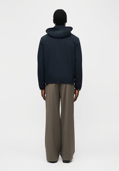 Άτομο που στέκεται με την πλάτη, φορώντας μαύρο μπουφάν με κουκούλα και φαρδιά χακί παντελόνια, πάνω σε απλό λευκό φόντο.