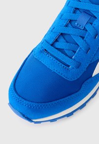 Sneaker atletica blu con tessuto testurizzato e sovrapposizioni in suede, lacci piatti e una suola intermedia bianca su uno sfondo grigio chiaro.