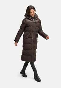 Brun puffermantel med huva, dragkedja och knappstängning, två framfickor och en lång, figursydd design. Modellen bär svarta ankelskor.