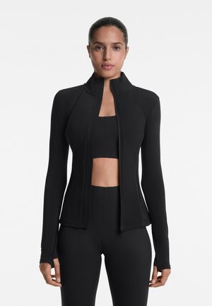Veste de sport noire à manches longues avec un col haut, fermeture éclair intégrale et design ajusté, assortie avec un crop top noir et un pantalon taille haute.