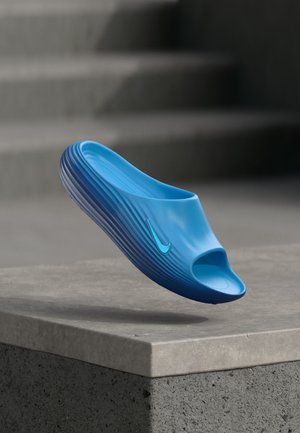 Μπλε σανδάλι Nike slide με ανοιχτή μύτη και υφή στη σόλα, αιωρούμενο πάνω από μια τσιμεντένια άκρη και σκάλες στο φόντο.