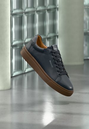 Black leather sneaker with brown sole and laces, suspended mid-air against a glossy glass block wall and concrete floor background.