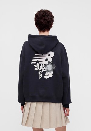 Personne aux cheveux courts et bouclés portant un sweat à capuche noir avec un motif floral blanc et le logo NB dans le dos, associé à une jupe plissée beige.
