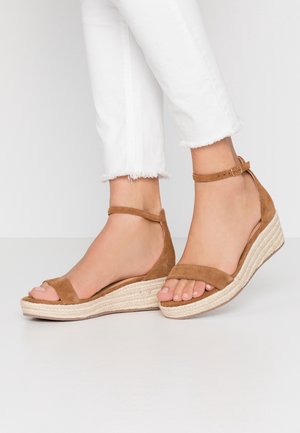 Voeten in tan kleur suede wedge sandalen met enkelriemen en witte broek met rafelige zoom tegen een effen achtergrond.