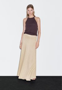 Woman standing against white wall wearing sleeveless dark brown top and long beige skirt, with neutral expression and hands relaxed by sides.