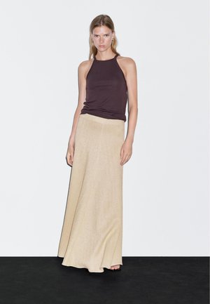 Vrouw staande tegen een witte muur, gekleed in een mouwloos donkerbruin topje en een lange beige rok, met een neutrale uitdrukking en handen ontspannen langs haar zij.