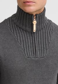 Grå strikket sweater med høj ribbet krave, halv-zip lukning, og et tekstureret mønster omkring hals og krop. Messing lynlås detalje synlig.