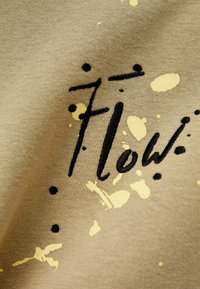 Beige tyg med svart broderad text "Flow" och gula färgsplashes. Nära bild visar textur och design detaljer.