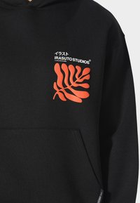 Černá mikina s kapucí a přední kapsou. Ozdobena jasně oranžovým designem listu a logem "IRASUTOSTUDIOS" v bílé barvě. Měkká textilie.