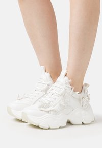 Witte sneakers van gestructureerd synthetisch materiaal, met reliefaccenten in de vorm van vlinders en een zware zool. Vetersluiting met gewatteerde kraag.
